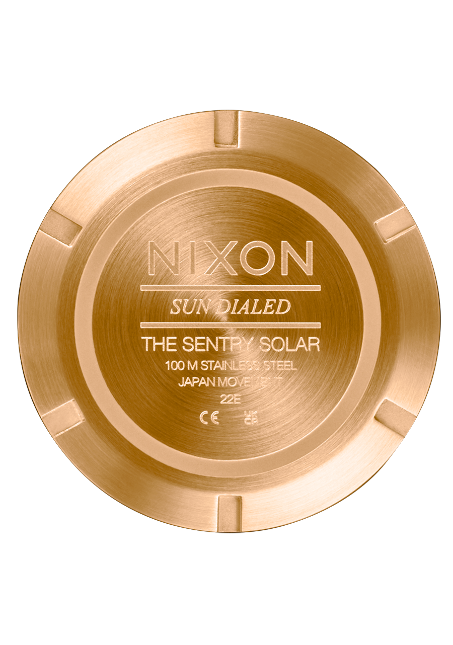 Gold-colored circular watch case back with ’Nixon’ branding and product details engraved.