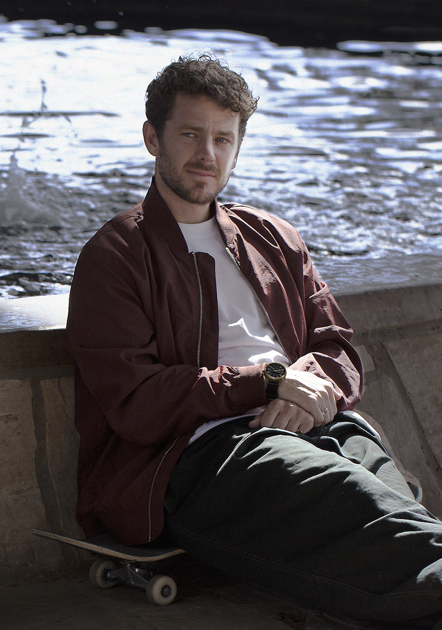 Man sitting on a skateboard wearing a maroon jacket and white shirt.