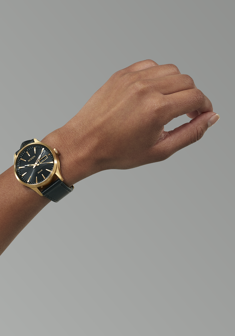 Gold-toned wristwatch with a black face and leather strap worn on a person’s wrist.