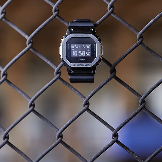 Digital wristwatch with a square face hanging on a chain-link fence.