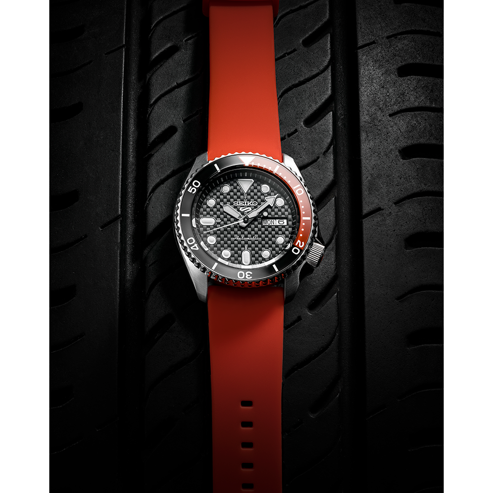 Wristwatch with a black dial and red strap against a tire tread background.