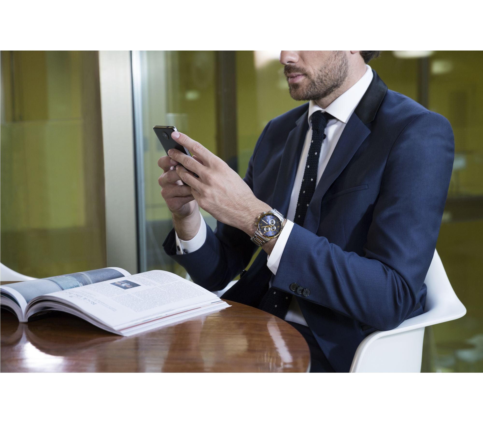 Businessman in a navy suit checking his smartphone while seated at a desk with an open book or document.